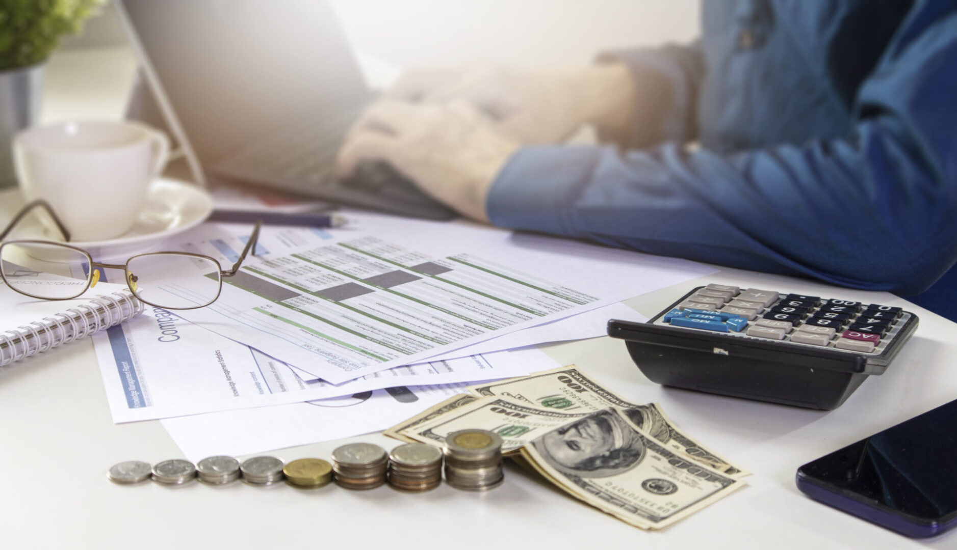 Calculator, money, and charts on a work desk with a person's hands in the background.